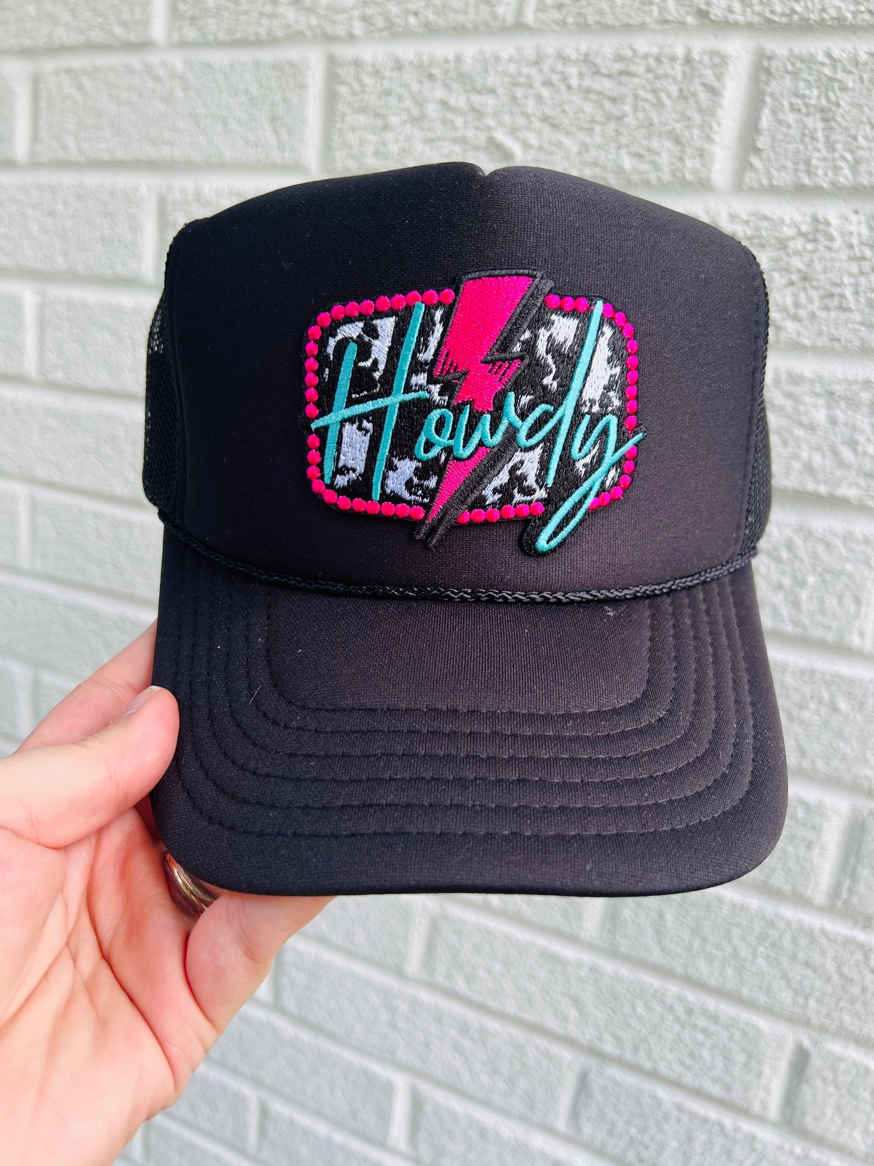 A black trucker hat with a colorful 'Howdy' text and lightning bolt embroidery patch on the front.
