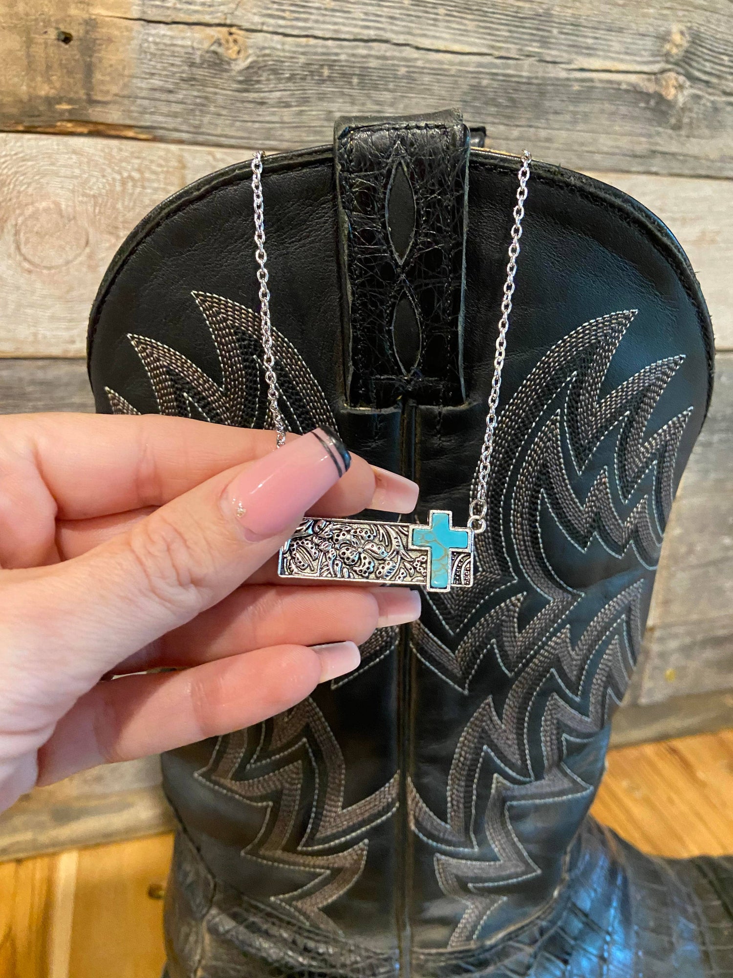 Hand holding a necklace with a cross pendant in front of a black boot with intricate designs.