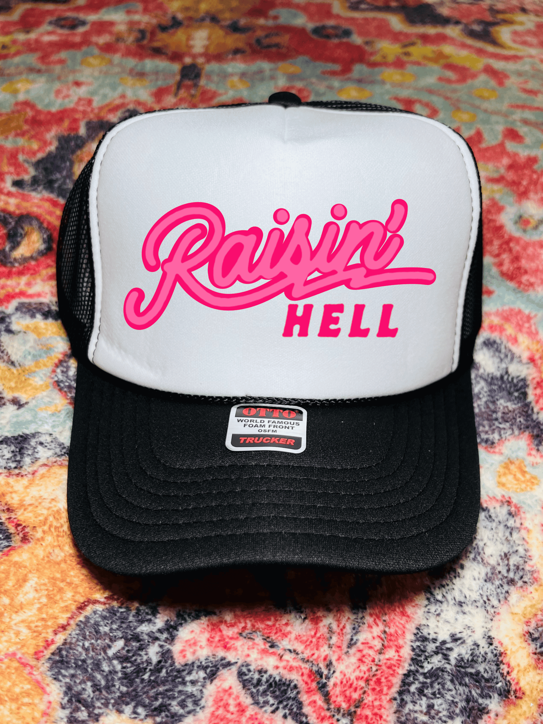 A black and white trucker hat with 'Raising Hell' text in neon pink on the front panel, displayed on a patterned background.