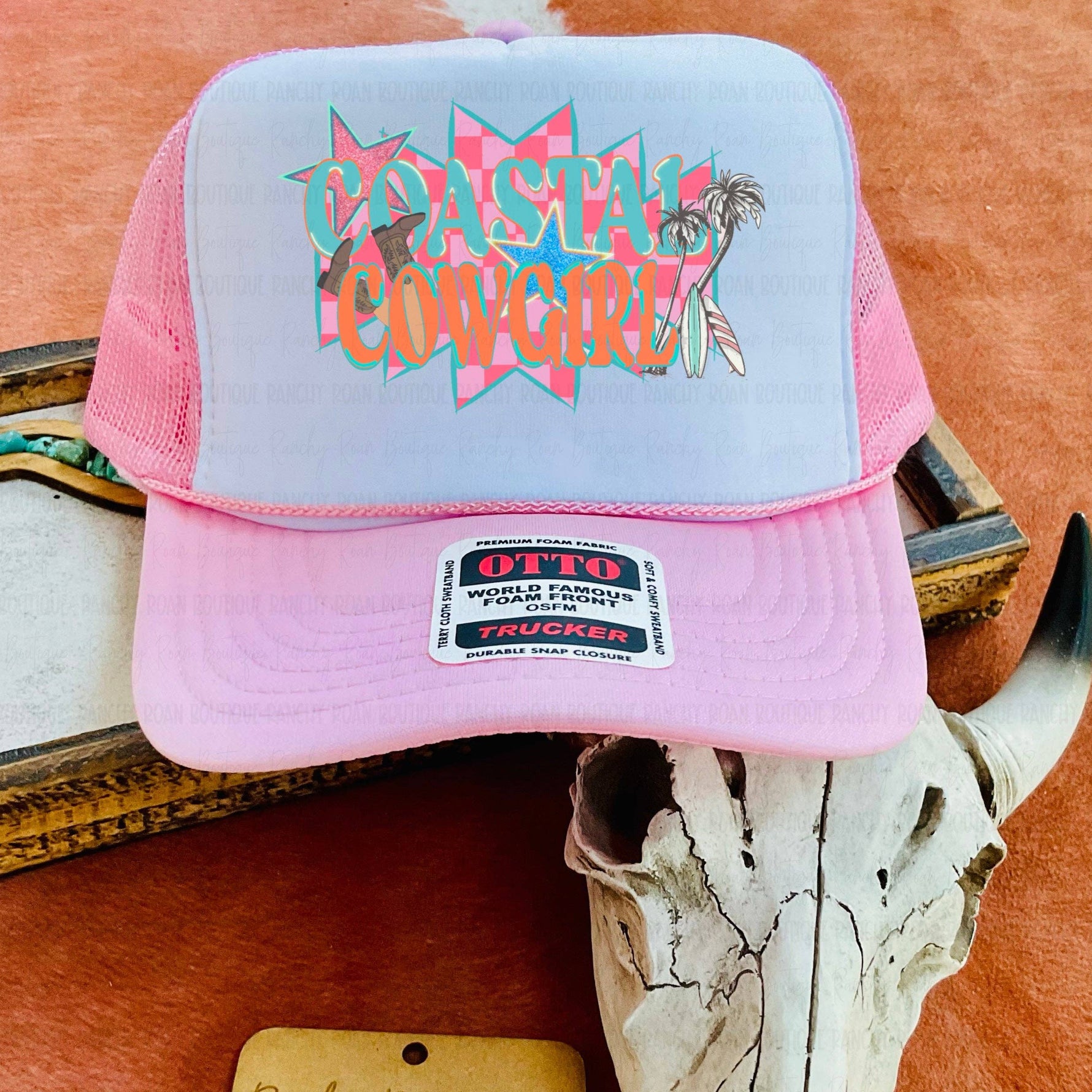 Pink and gray cap with 'Coastal Cowgirl' text on a wooden surface with a cow skull and wooden sign.