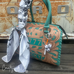 A turquoise and tan tooled leather handbag with a matching handle, decorated with playing card and dice charms, and styled with a white patterned scarf, set against a rustic metal background.