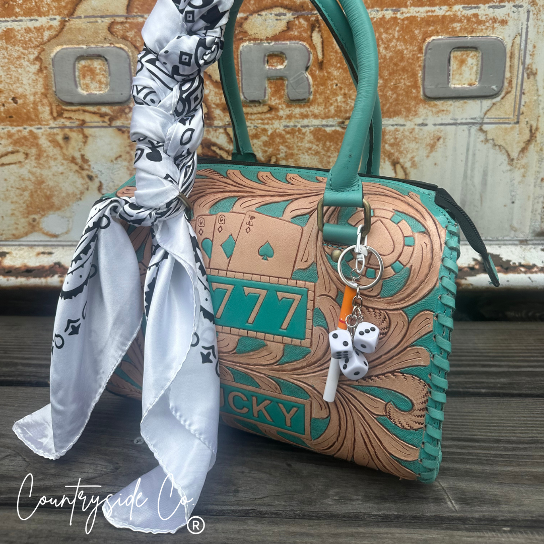 A turquoise and tan tooled leather handbag with a matching handle, decorated with playing card and dice charms, and styled with a white patterned scarf, set against a rustic metal background.