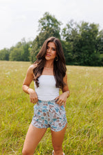 Woman standing in a field wearing a white top and patterned shorts.