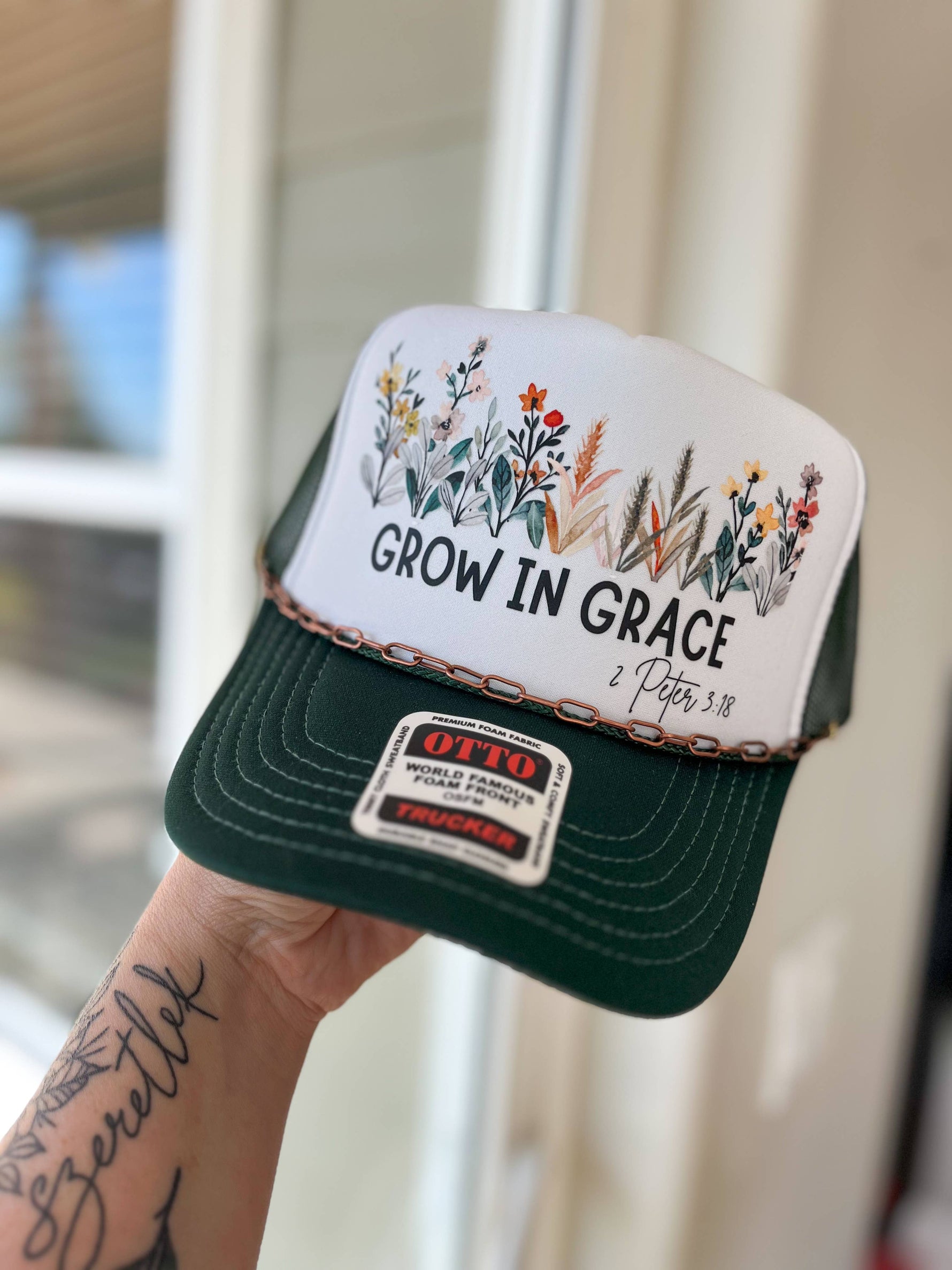 Green and white cap with 'Grow in Grace' text and floral designs, held by a person with tattoos.