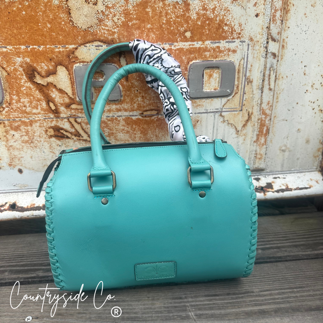 A turquoise leather handbag with braided trim and a black-and-white patterned scarf tucked into the handle, set against a rusted metal door background. A 'Countryside Co' watermark is visible in the bottom left corner.