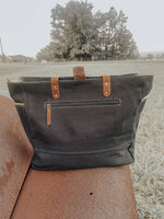 Black tote bag with brown straps on a stone surface with a blurred outdoor background