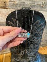 Hand holding a necklace with a cross pendant in front of a black boot with intricate designs.