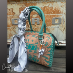 A turquoise and tan handbag with a matching turquoise handle, decorated with a white and black patterned scarf, dice charms, and a keychain, set against a rustic metal background.