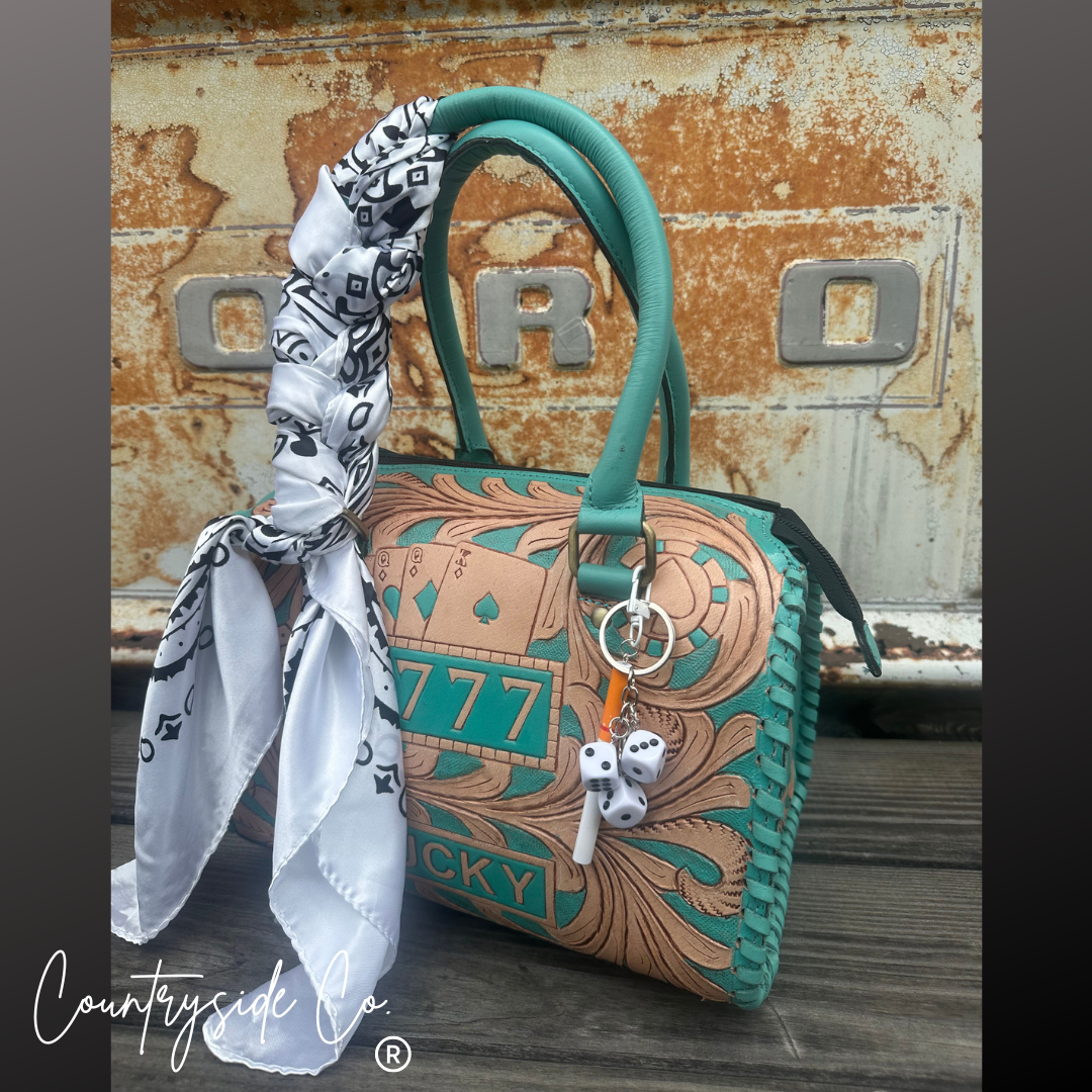 A turquoise and tan handbag with a matching turquoise handle, decorated with a white and black patterned scarf, dice charms, and a keychain, set against a rustic metal background.