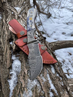 Folded knife with a detailed blade and red sheath attached to a tree in a snowy setting