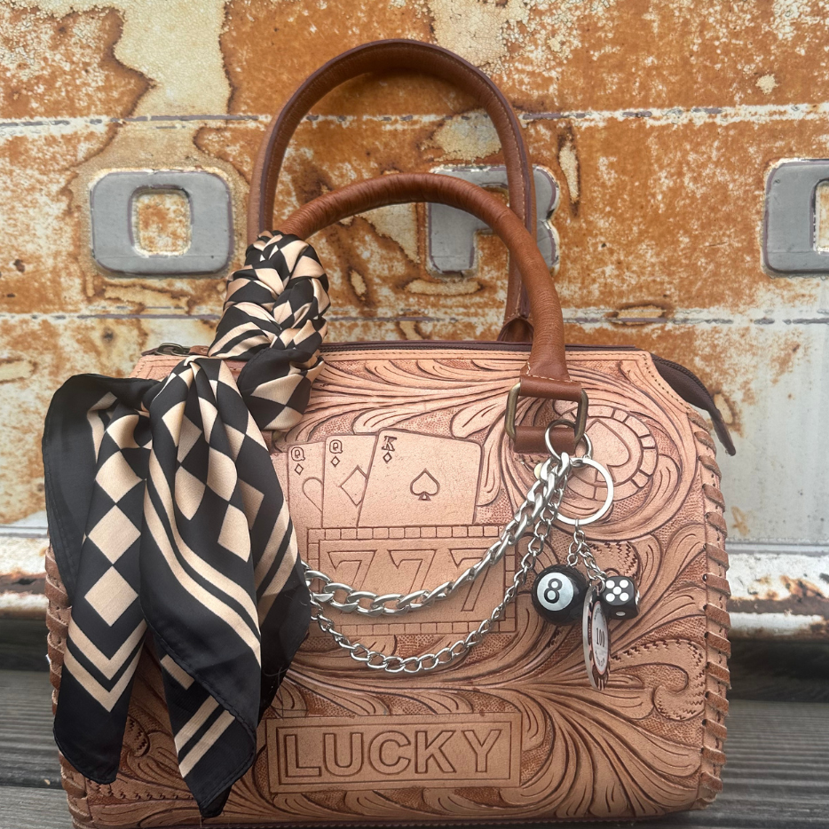 A tooled brown leather handbag with embossed playing card designs and the word 'LUCKY' on the front, styled with a black and beige patterned scarf and silver chain charms. The bag is photographed outdoors against a rustic, rusted metal surface.