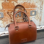 A brown leather handbag with double top handles and a patterned fabric loop at the top, resting on a weathered wooden surface against a rusted metal door background. 