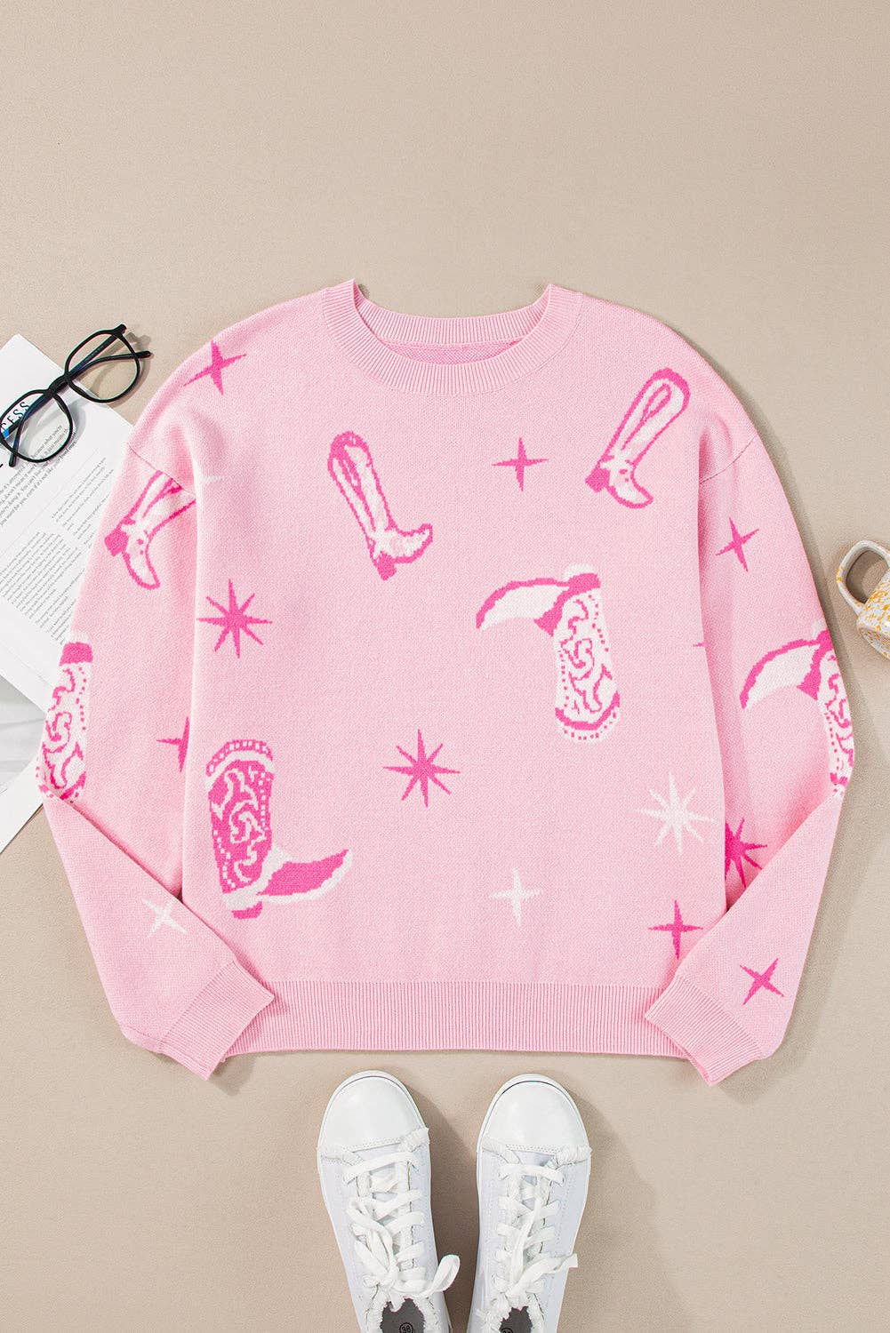 Pink sweatshirt with graphic designs on a beige surface with white sneakers and glasses.