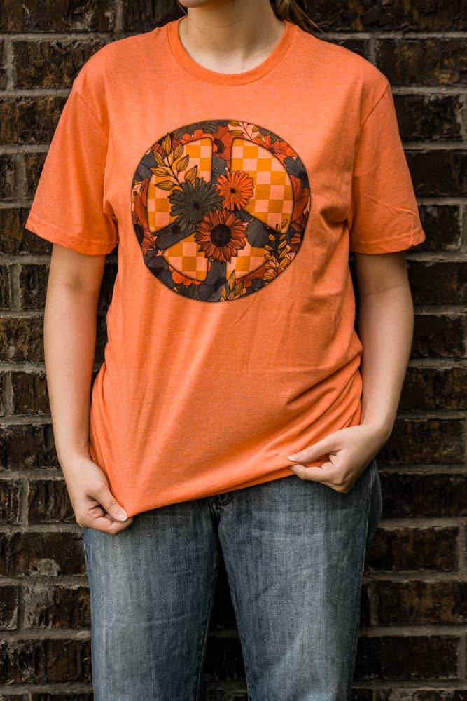 Person wearing an orange t-shirt with a floral peace symbol design against a brick wall.