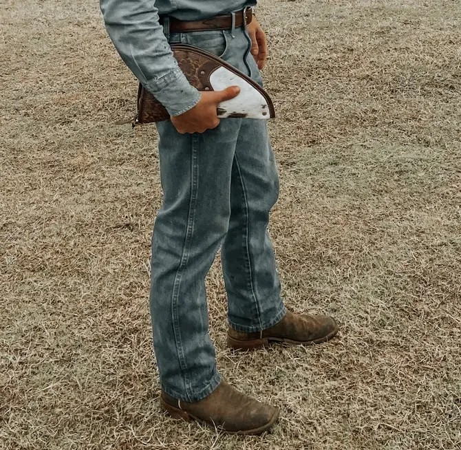 Person wearing blue jeans and brown boots holding a leather jacket in a field.
