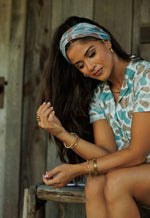 Woman sitting outdoors wearing a patterned headscarf and shirt.