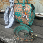 A turquoise and tan tooled leather handbag with a matching strap, displayed on a weathered wooden surface. The bag features a teal and tan tooled leather body with a '777 LUCKY' design, a white bandana tied to the handle, and a lighted cigarette charm.