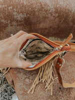 Hand holding a brown leather bag with fringe against a rustic background