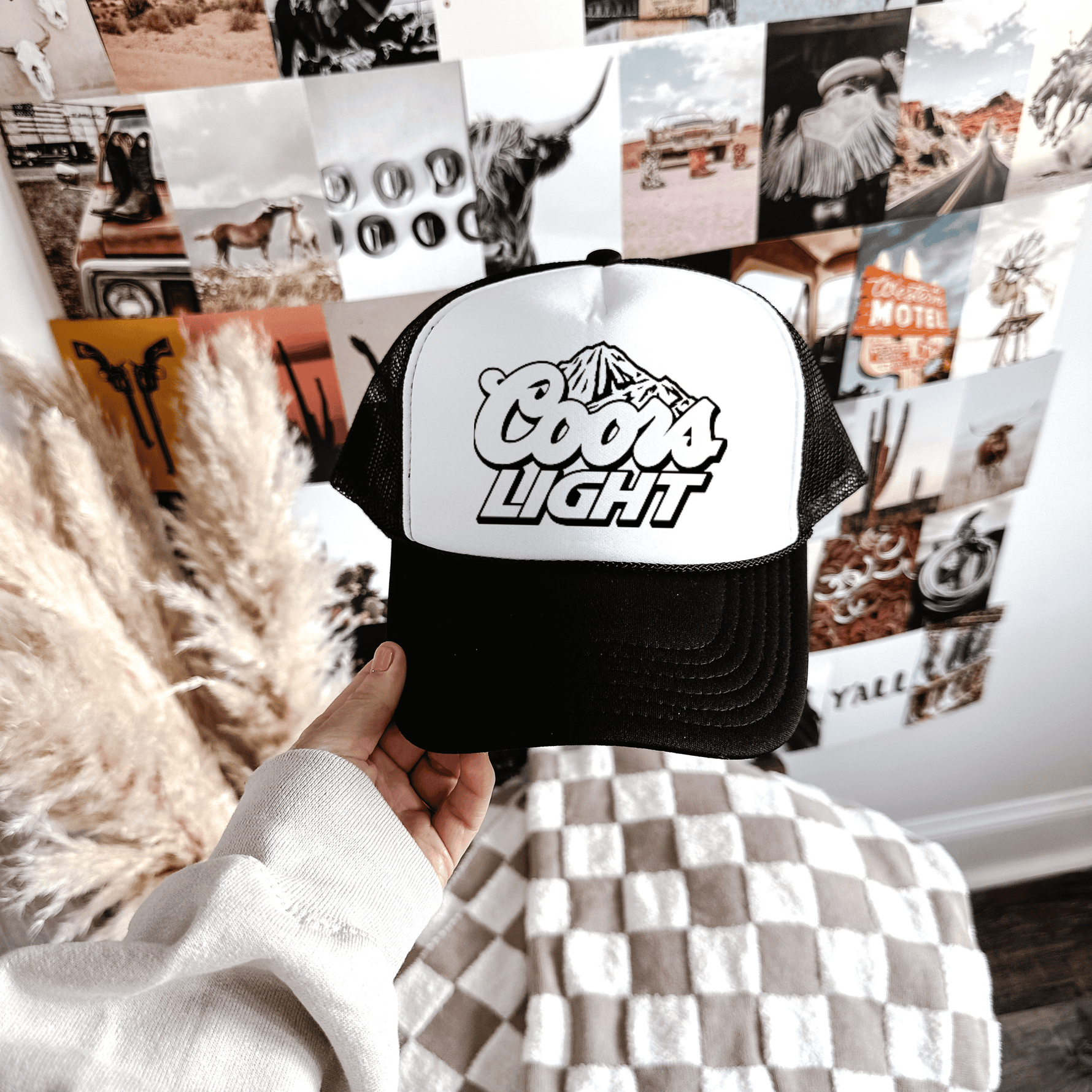 A hand holding a black and white Coors Light trucker hat with a checkered pattern on the visor, displayed against a backdrop with various photos and a decorative element.