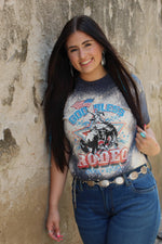 A woman with long dark hair stands against a textured concrete wall wearing a faded blue graphic t-shirt with a cowboy riding a bucking bull design and the words 'God Bless Rodeo'. She is also wearing blue jeans with a silver concho belt.