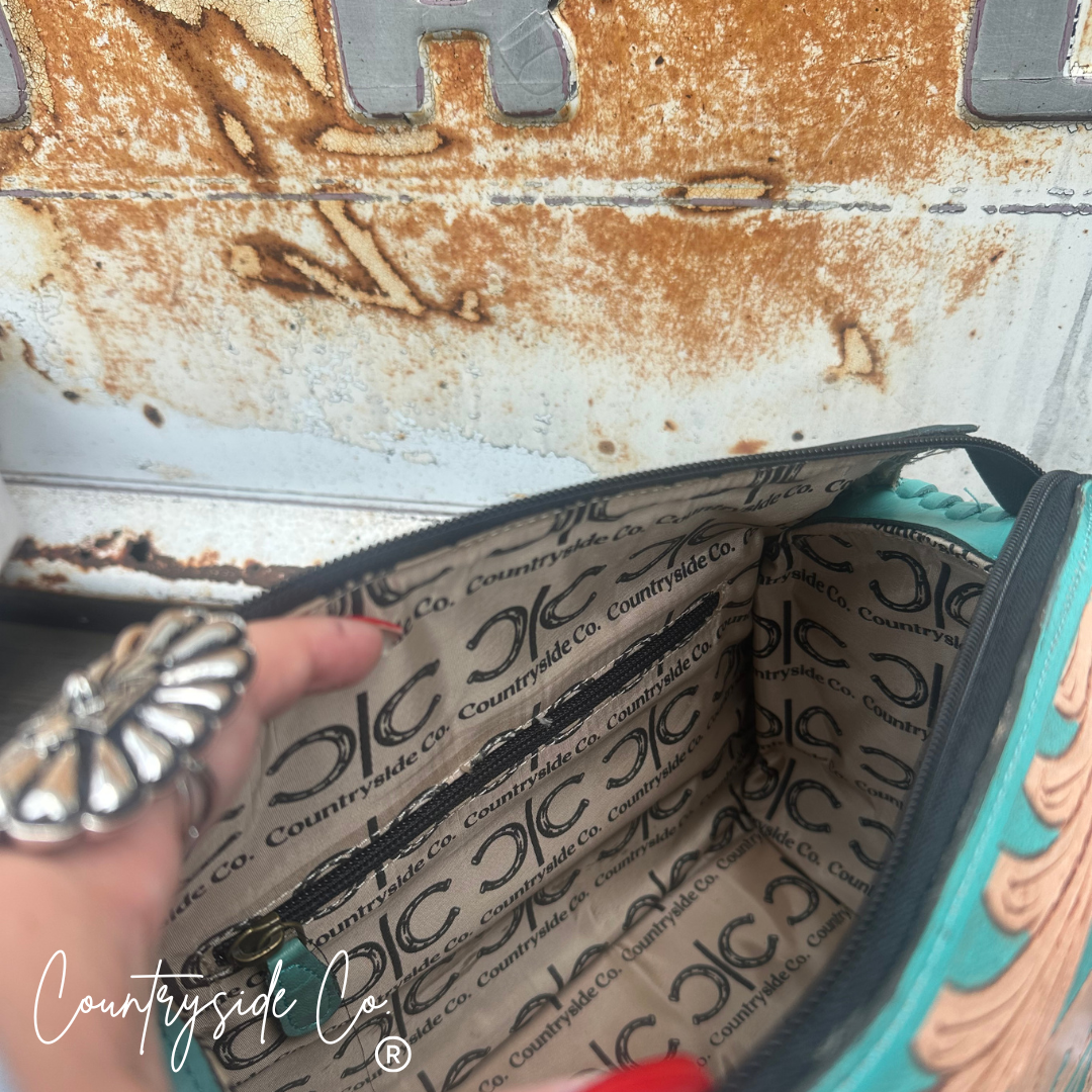 An open turquoise and beige patterned zippered pouch or cosmetic bag, shown with a hand inside. The interior lining has a repeating black 'Countryside Co' logo pattern.