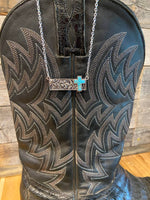 Black leather boot with intricate stitching and a silver necklace with a turquoise cross on a wooden surface.