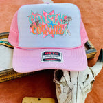 Pink and gray cap with 'Coastal Cowgirl' text on a wooden surface with a cow skull and wooden sign.